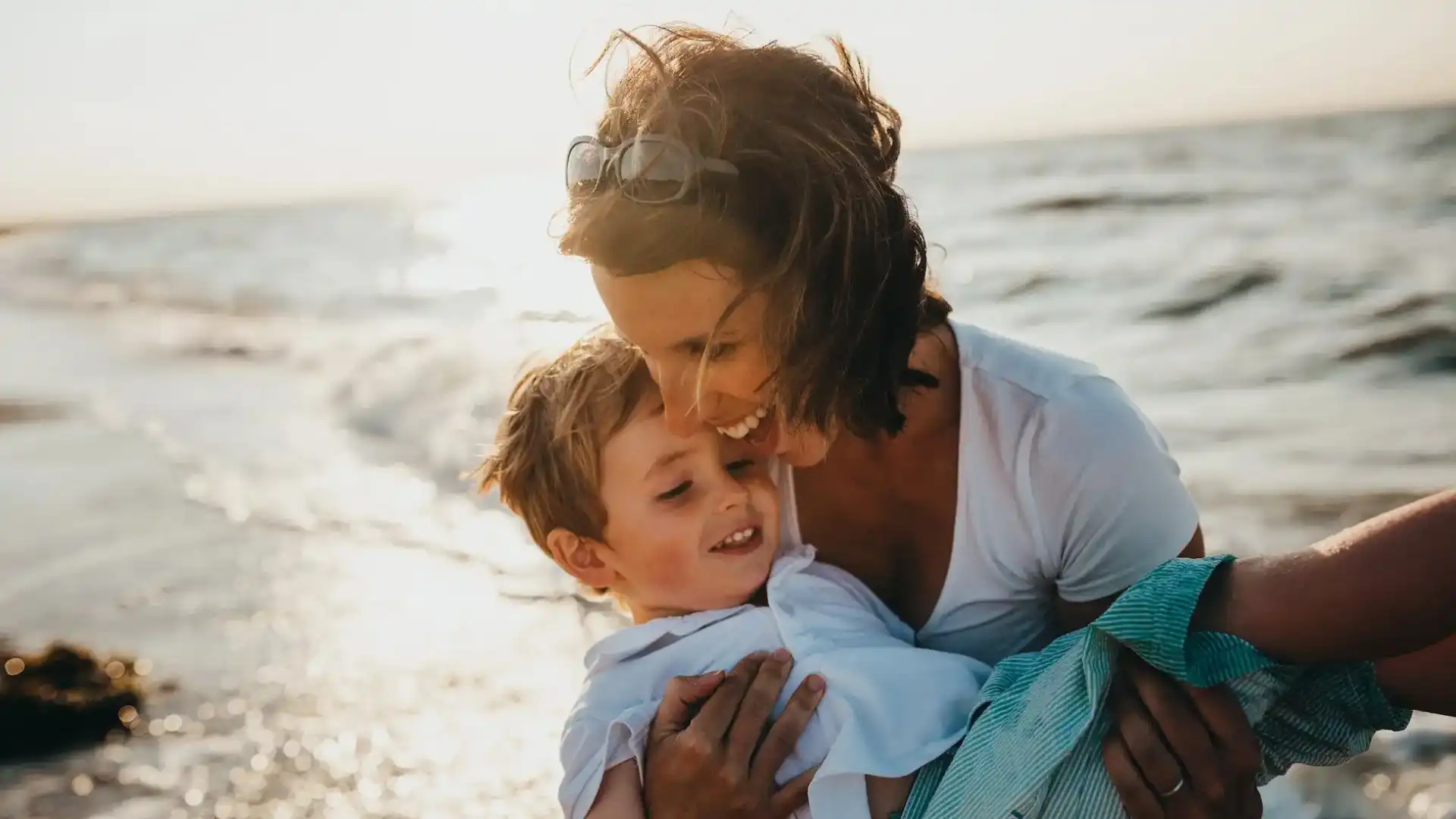 Uma mãe sorridente segura seu filho na beira da praia, enquanto a luz do sol brilha ao fundo, criando um momento especial entre eles.