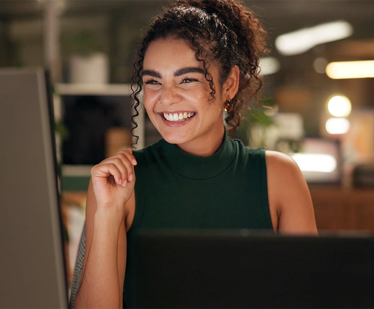 Mulher sorrindo e feliz enquanto trabalha no computador, cenário de escritório acolhedor com iluminação quente e plantas ao fundo.