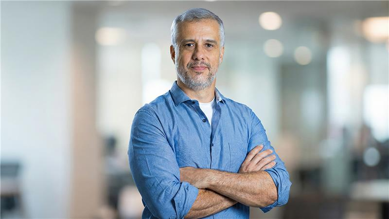 Homem de meia-idade com barba grisalha, vestindo camisa jeans, cruzando os braços em ambiente de escritório moderno, transmitindo confiança e experiência.