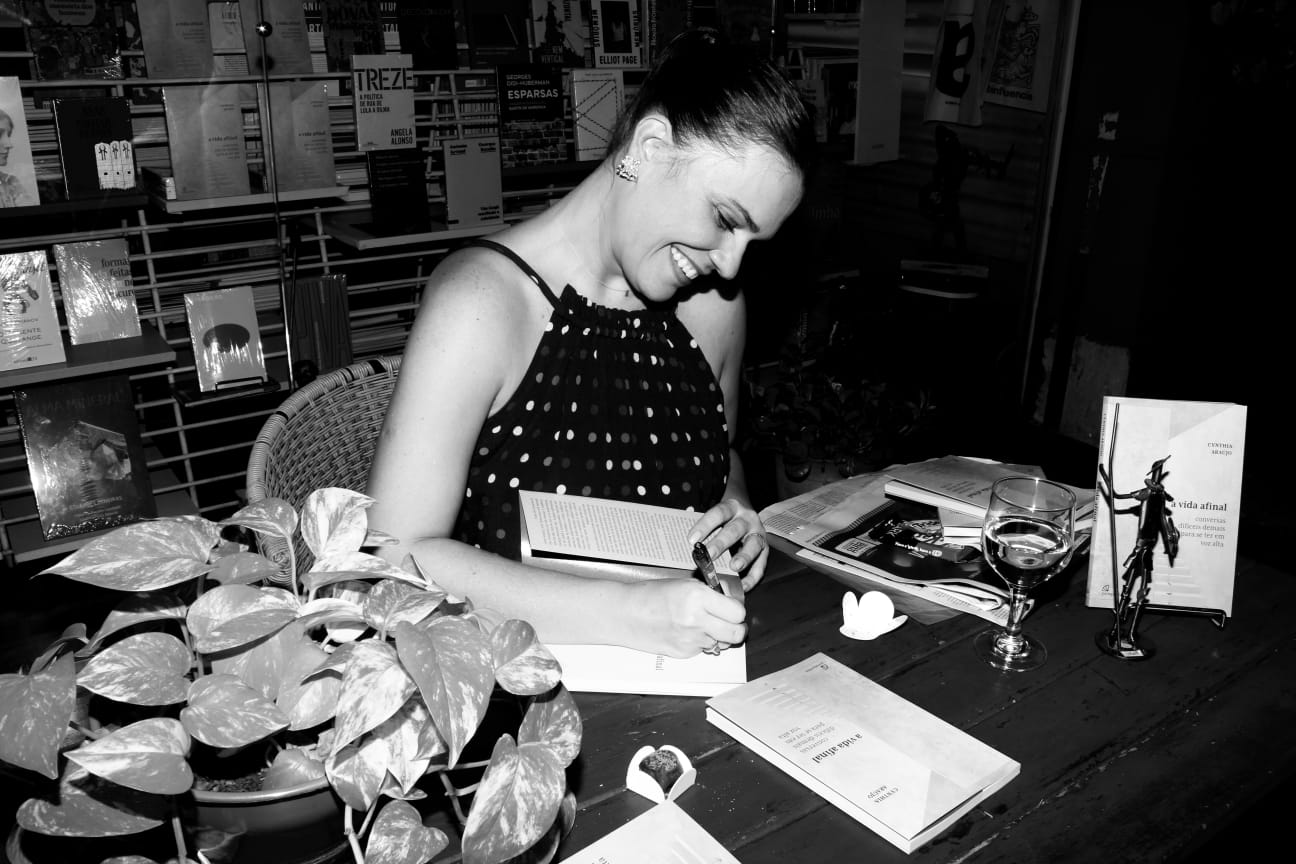 Mulher sorridente lendo um livro em uma livraria, cercada por revistas e uma planta, com uma taça de vinho ao lado. Ambiente aconchegante e tranquilo.