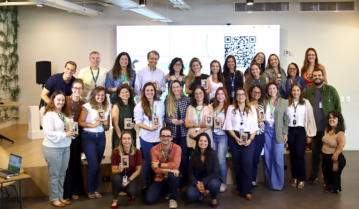 Grupo de pessoas em uma conferência ou evento corporativo, segurando certificados ou cartões de identificação, em ambiente moderno com grande tela de QR code ao fundo.