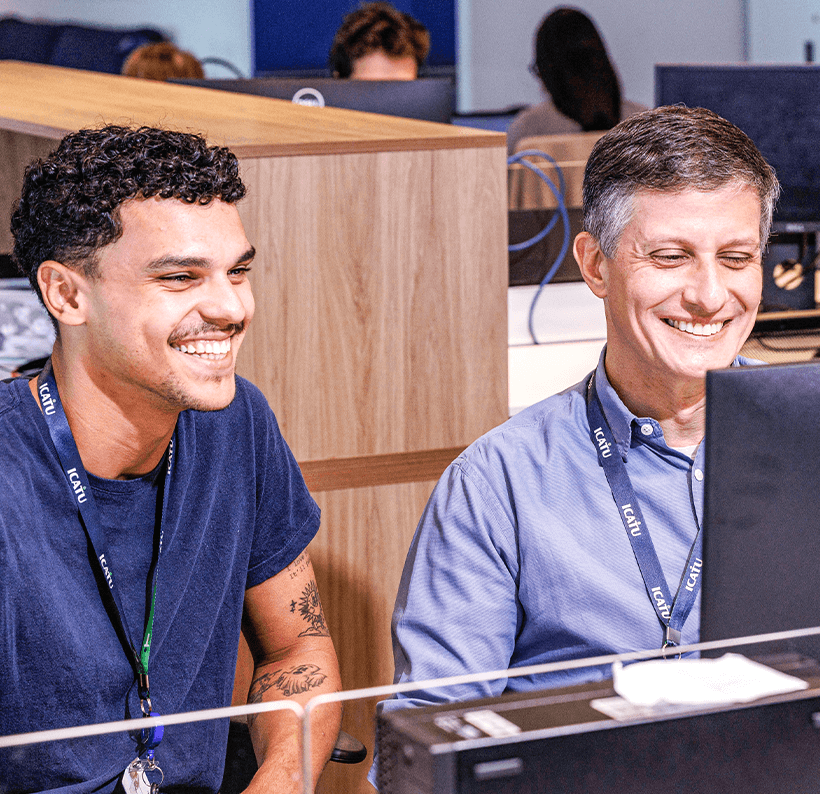 Homens sorrindo em ambiente de escritório com computadores, participando de evento de tecnologia ou treinamento, ambiente profissional e descontraído.